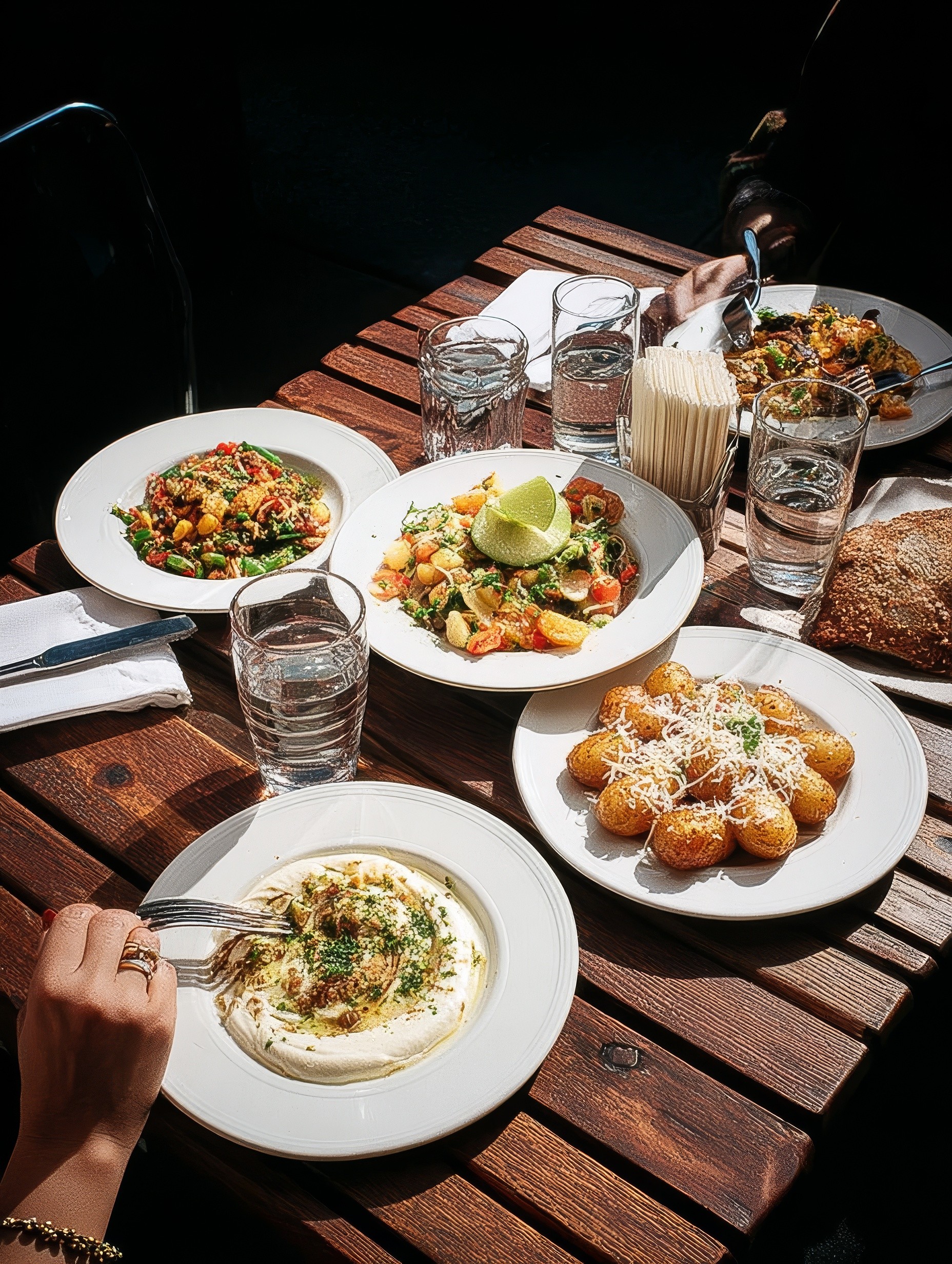 Wooden outdoor table set with multiple plates of food, including dips, salads, potatoes with cheese, and glasses of water.