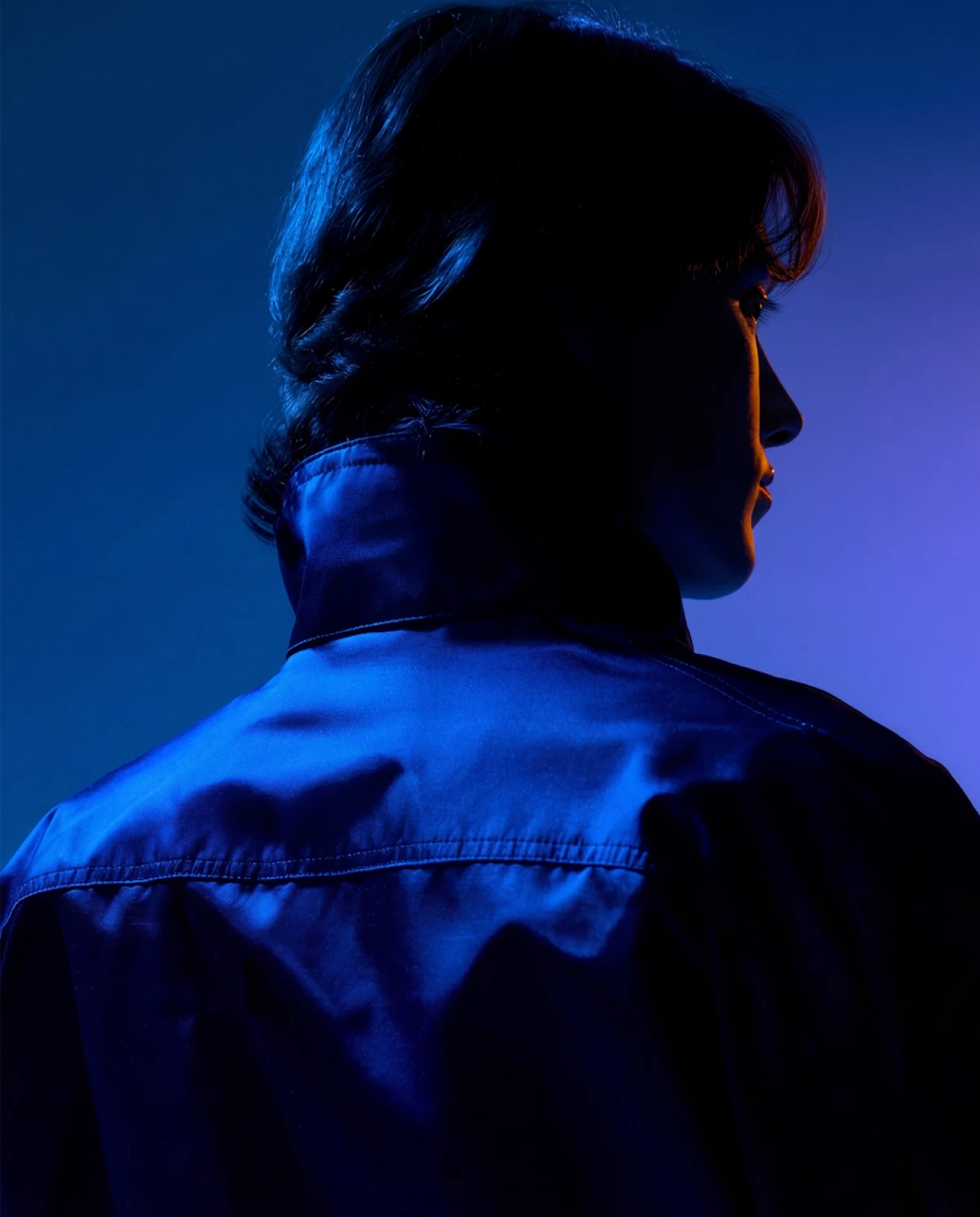 Profile of a person with medium-length hair wearing a dark blue jacket, illuminated by blue and purple lighting.