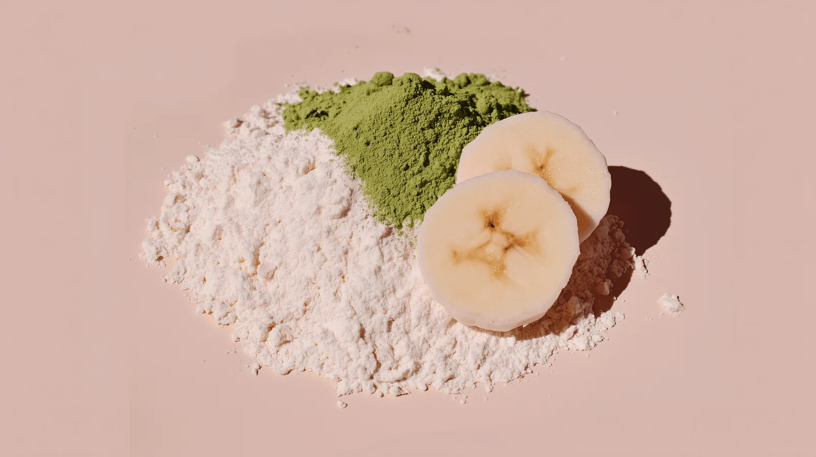 Matcha powder, protein powder and banana slices arranged on a blush pink background